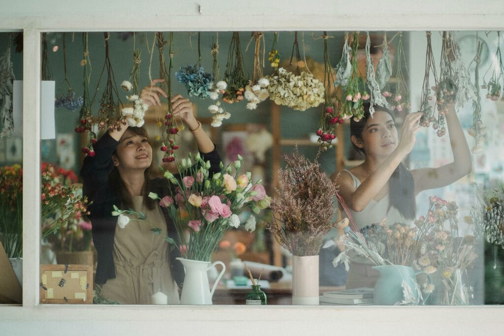 Through window content multiracial female floral shop workers wearing aprons decorating store window with delicate flowers